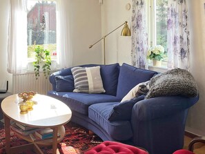 A living room with a blue sofa, a round table with books, a floor lamp, and a window with curtains. - 6 Person Holiday Home in Klintehamn (Klintehamn)