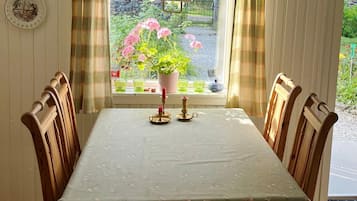 A dining table with a floral tablecloth, surrounded by wooden chairs, with a window view of a garden and a potted plant.