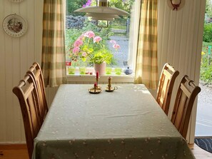 Une table à manger recouverte d’une nappe à motifs floraux, entourée de chaises en bois, avec une fenêtre donnant sur un jardin et une plante en pot.