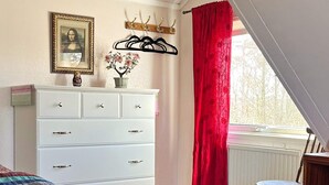 Ein Schlafzimmer mit einem weißen Schminktisch, einem Bett mit bunter Steppdecke, einem roten Vorhang und einem Fenster mit Blick.