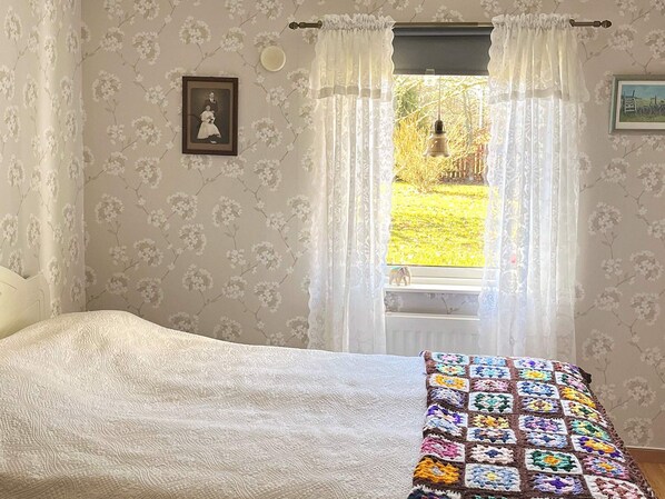 Une chambre à coucher avec un lit, une fenêtre avec des rideaux, un papier peint à motifs floraux et un tableau encadré accroché au mur.