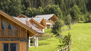 Une rangée de cabanes en bois, posées sur des fondations en pierre, se trouve sur une colline verdoyante, avec une clôture en bois en premier plan.