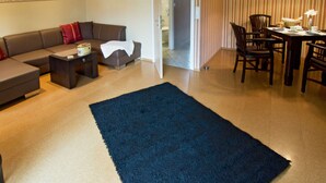 Una sala de estar con sofá, mesa de centro y alfombra azul. Se observa una zona de comedor con mesa y sillas. En la pared, se ve un reloj.