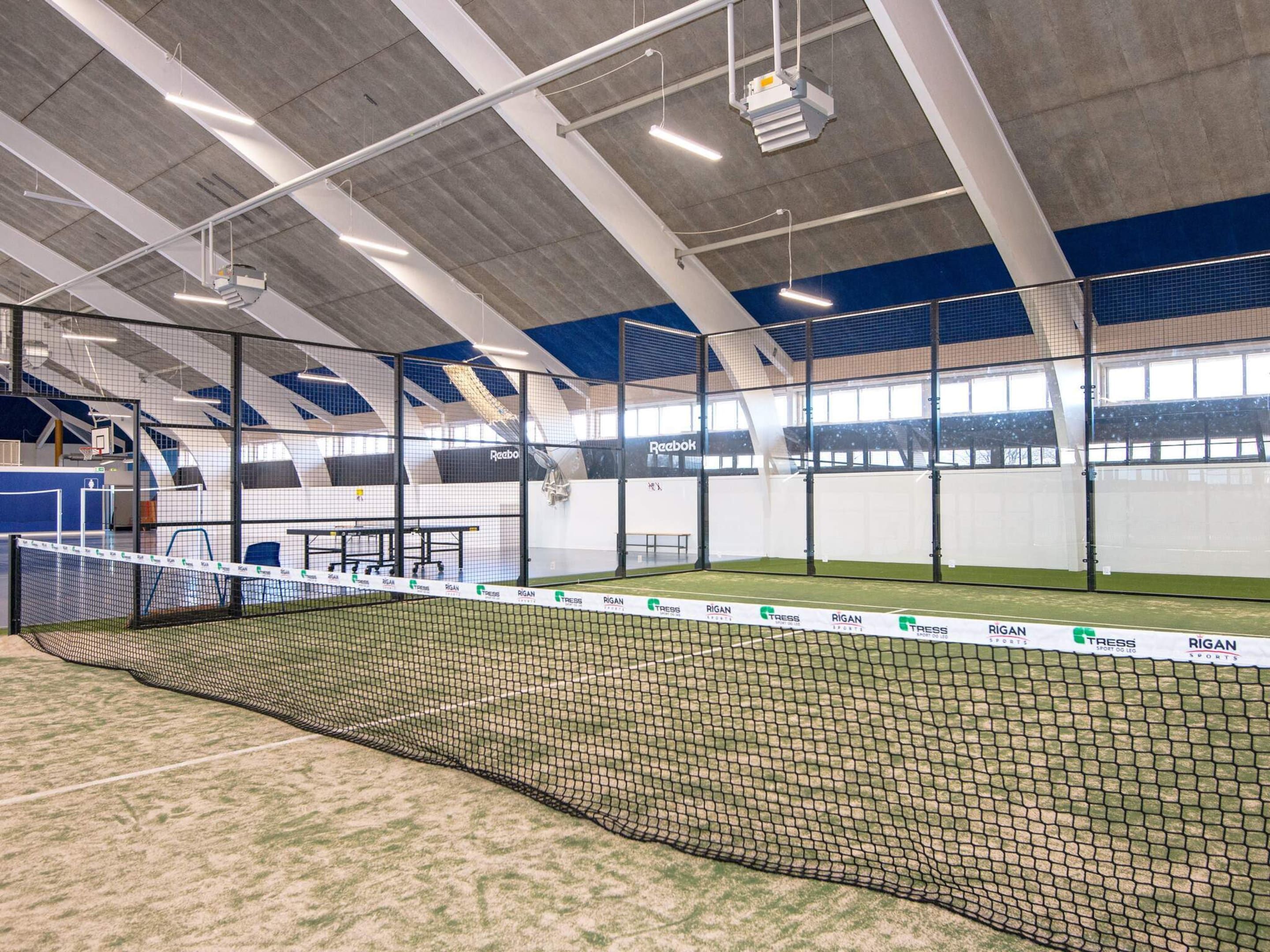 An indoor sports facility with a tennis court, seating areas, and a high ceiling with lighting fixtures.
