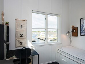 Une pièce avec une fenêtre, un bureau, une chaise, un lit et une étagère fixée au mur, sur laquelle se trouve une lampe.