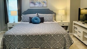Une chambre à coucher bien rangée, avec une couvre-lit à motifs, une tête de lit, une table de chevet avec une lampe, un téléviseur posé sur un meuble et un tableau accroché au mur.