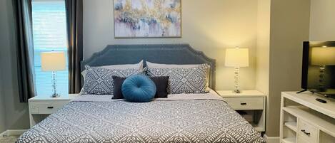 Un dormitorio ordenado con una colcha estampada, un cabecero, una mesita de noche con lámpara, un televisor sobre un mueble y un cuadro en la pared.