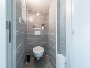 A modern bathroom with grey tiles, a white toilet, a small shelf with decorative items, and a toilet paper holder. - Studio on the 3rd Floor With Phenomenal sea View (Middelkerke)