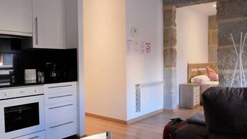 Cozinha moderna com armários brancos, bancada preta e forno embutido. Na sala adjacente, uma parede de pedra com radiador e uma cama com cabeceira de madeira.