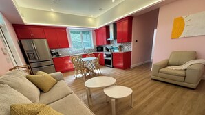 A modern living room with a beige sofa, two white round tables, and a beige armchair. A red kitchen with a large window and a dining area with wooden chairs is visible in the background.