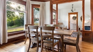 A dining area with a wooden table and chairs, large windows with curtains, and a view of greenery outside.