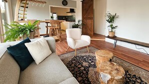 A modern living room with a sofa, a white armchair, a wooden coffee table, and a patterned rug. There is a kitchen in the background with a wooden door and a plant on the windowsill.