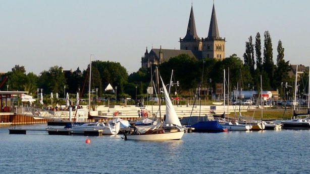 Een jachthaven met aangemeerde boten, een kerk met twee torens in de verte, en een waterkantpromenade met bomen en gebouwen.
