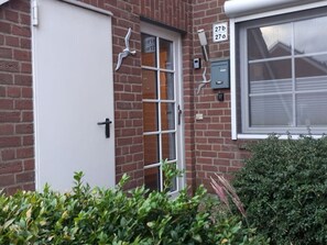 A brick building with a white door and window, a mailbox, and a small bush in the foreground. - Holiday Apartment Meerzeit (Brodersby)