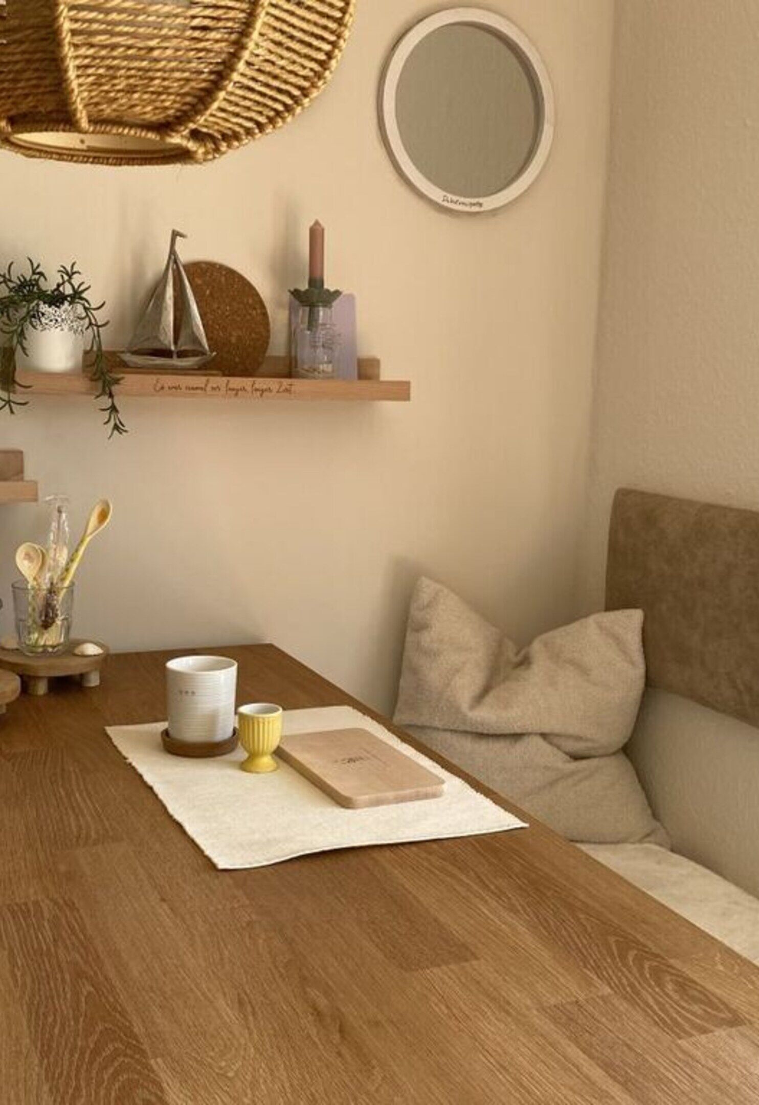 A cozy living room with a wooden table, a woven chair, a round mirror, and a beige sofa with cushions.