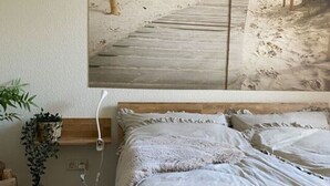 A bedroom with a bed, a wall-mounted painting of a beach, a bedside plant, and a wooden nightstand. - Holiday Apartment Meerzeit (Brodersby)