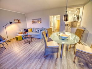 Una sala moderna con un sofá azul, sillas de mimbre, una mesa de comedor de vidrio y una cocina al fondo.