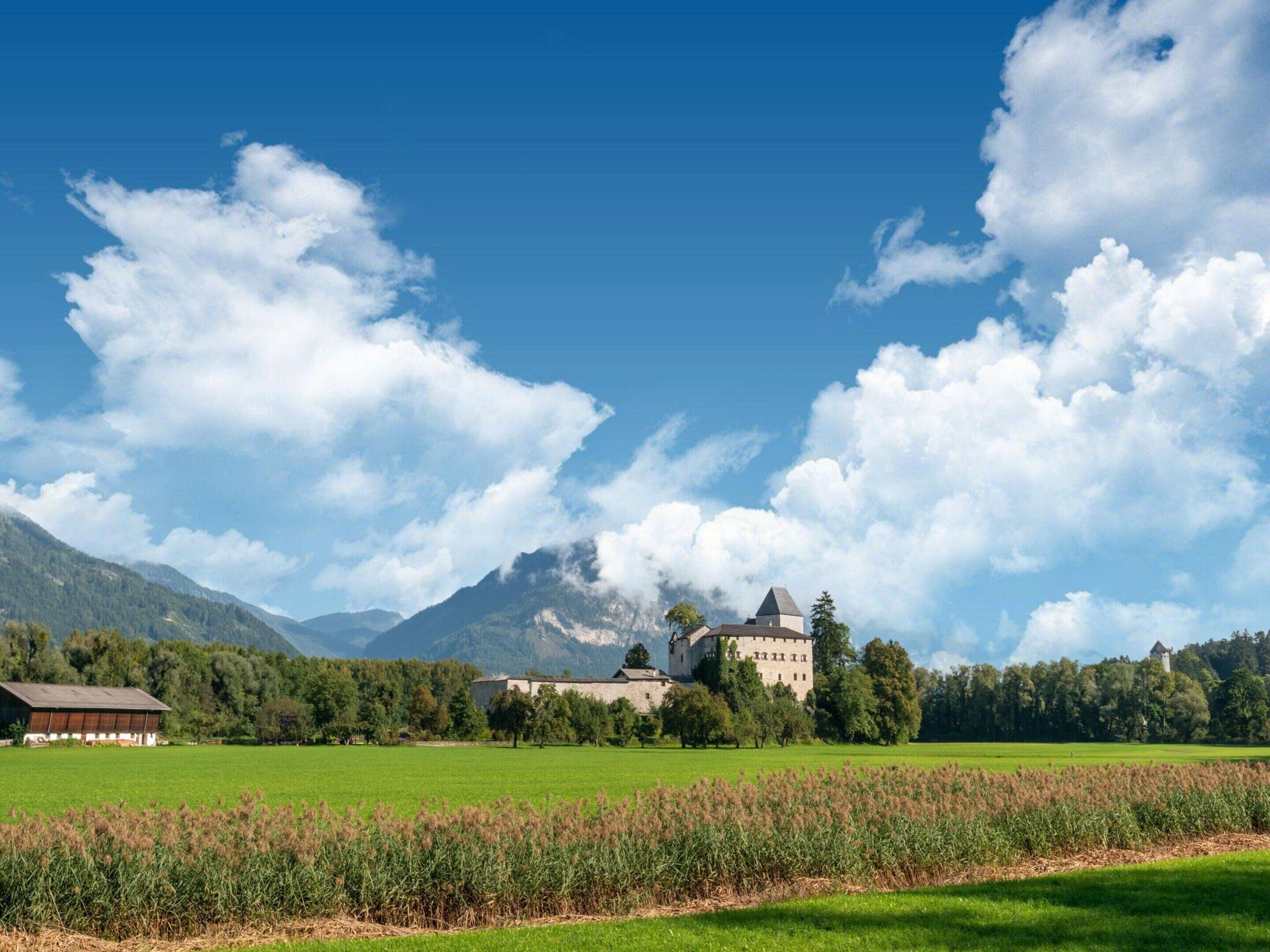A castle with a tower, surrounded by green fields and mountains under a clear sky.