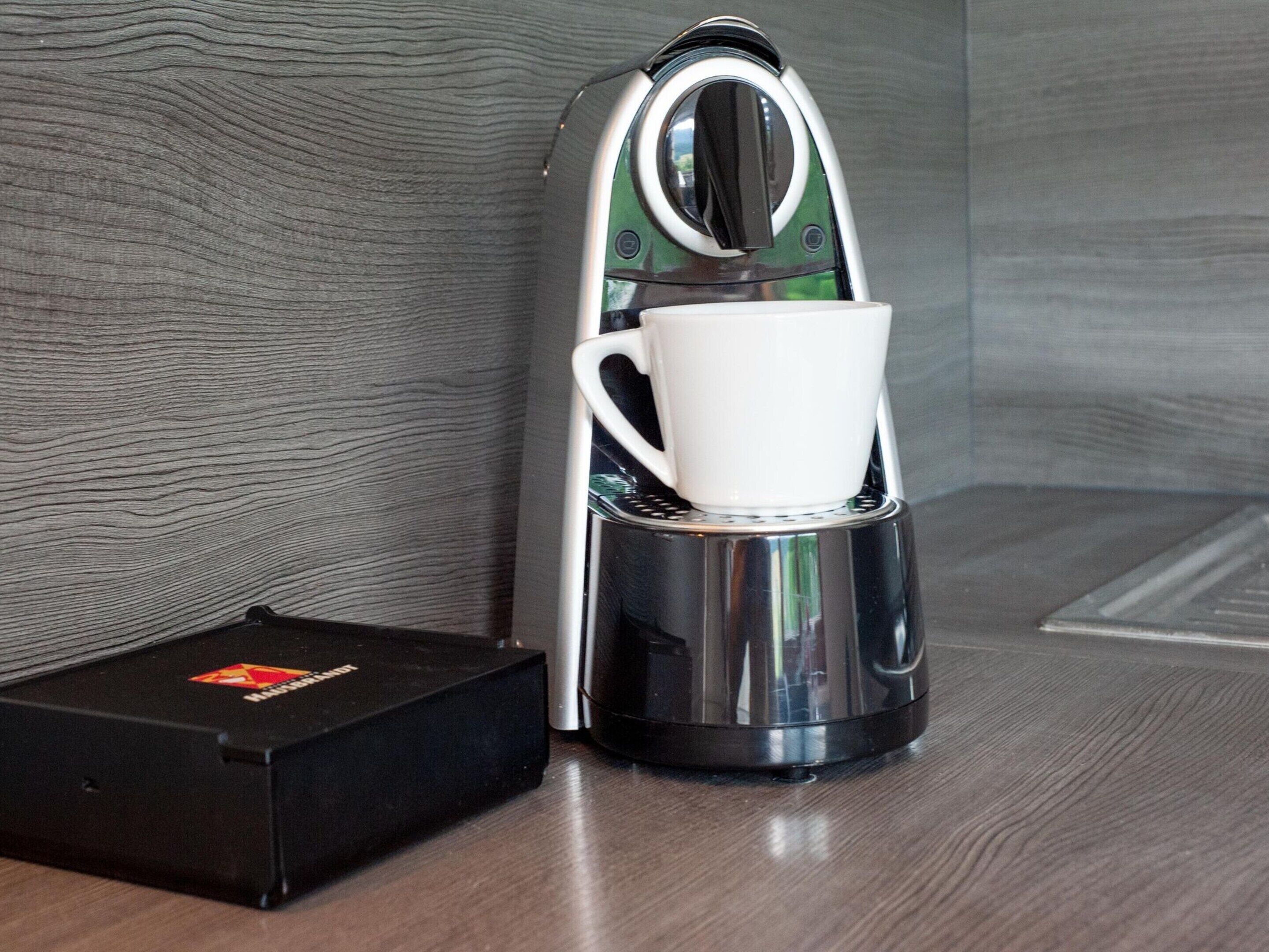 A coffee maker with a cup on its platform, placed on a wooden surface next to a black box.