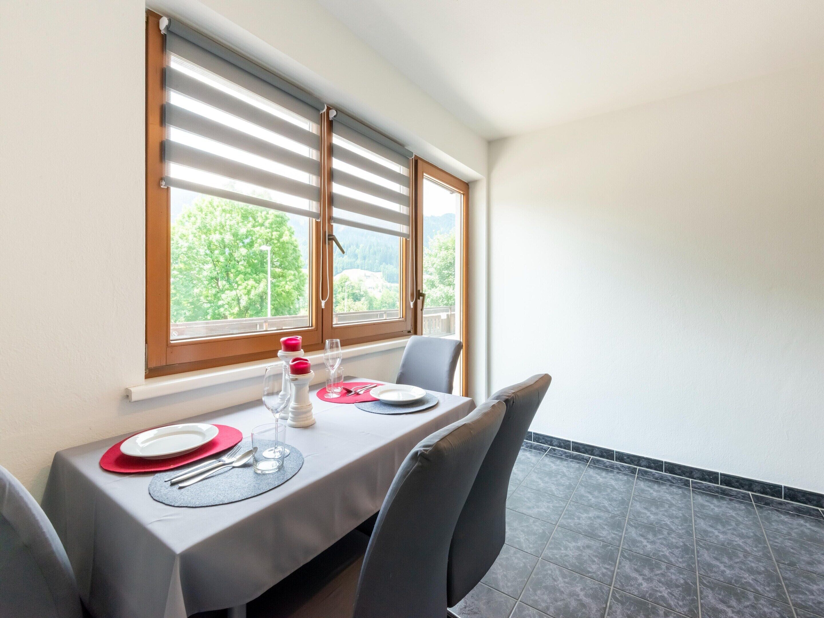 Comedor con una mesa preparada para dos personas, dos ventanas con persianas y vistas a zonas verdes.