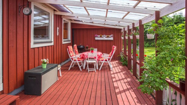 A covered outdoor patio with a wooden deck, a table with chairs, and a small garden area. - Nyhyttan Comfortable Holiday Residence (Nora)