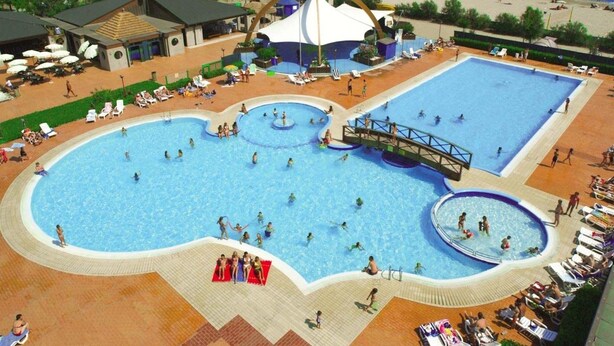 A large outdoor swimming pool with multiple sections, surrounded by lounge chairs and umbrellas, with a beach and ocean view in the background. - Apartment in Caorle Near the Beach (Caorle)