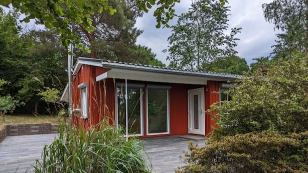 A red single-story house with a wooden deck, large sliding glass doors, and a garden area with various plants. - Holiday Home Werder/havel (Werder (Havel))