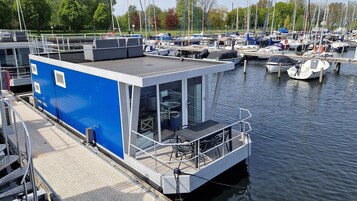 Una estructura azul flotante con terraza cubierta, ventanas y un rincón con mesa y sillones.