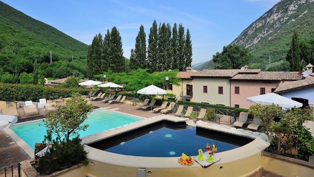 A pool area with lounge chairs and umbrellas, surrounded by greenery and buildings. - Apartment in Umbria With Pool and Sauna (Foligno)