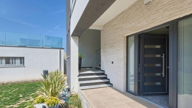 Un bâtiment moderne doté d’une porte en verre, d’un escalier menant à une entrée abritée et d’un petit jardin agrémenté de plantes.