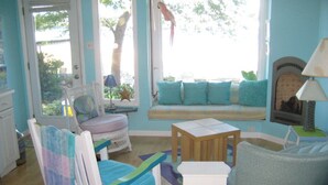 Una habitación acogedora con cama, una silla, una mesita y un sofá con cojines. Se ve una ventana con vistas a árboles y un adorno colgante.