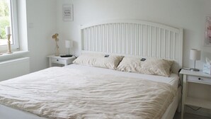 Cama bem arrumada com cabeceira branca e duas mesas de cabeceira, cada uma com uma luminária e um objeto decorativo.