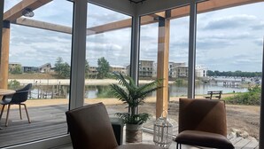 Ein modernes Zimmer mit großen Fenstern, einem Holzbalkon und Blick auf ein Gewässer sowie Gebäude.