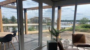 Ein moderner Außenbereich zum Sitzen mit einer Holzterrasse, einem Tisch mit Stühlen und Blick auf einen See sowie Gebäude in der Ferne.