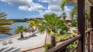 A beach view with palm trees, a pool, and a thatched-roof structure. - Holiday Home by Sea Willemstad Pool (Jan Thiel)