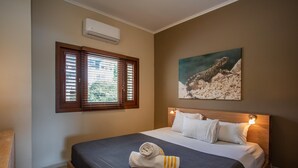 A hotel room with a bed, a window with blinds, a wall-mounted air conditioner, and a painting of a lizard. - Holiday Home by Sea Willemstad Pool (Jan Thiel)