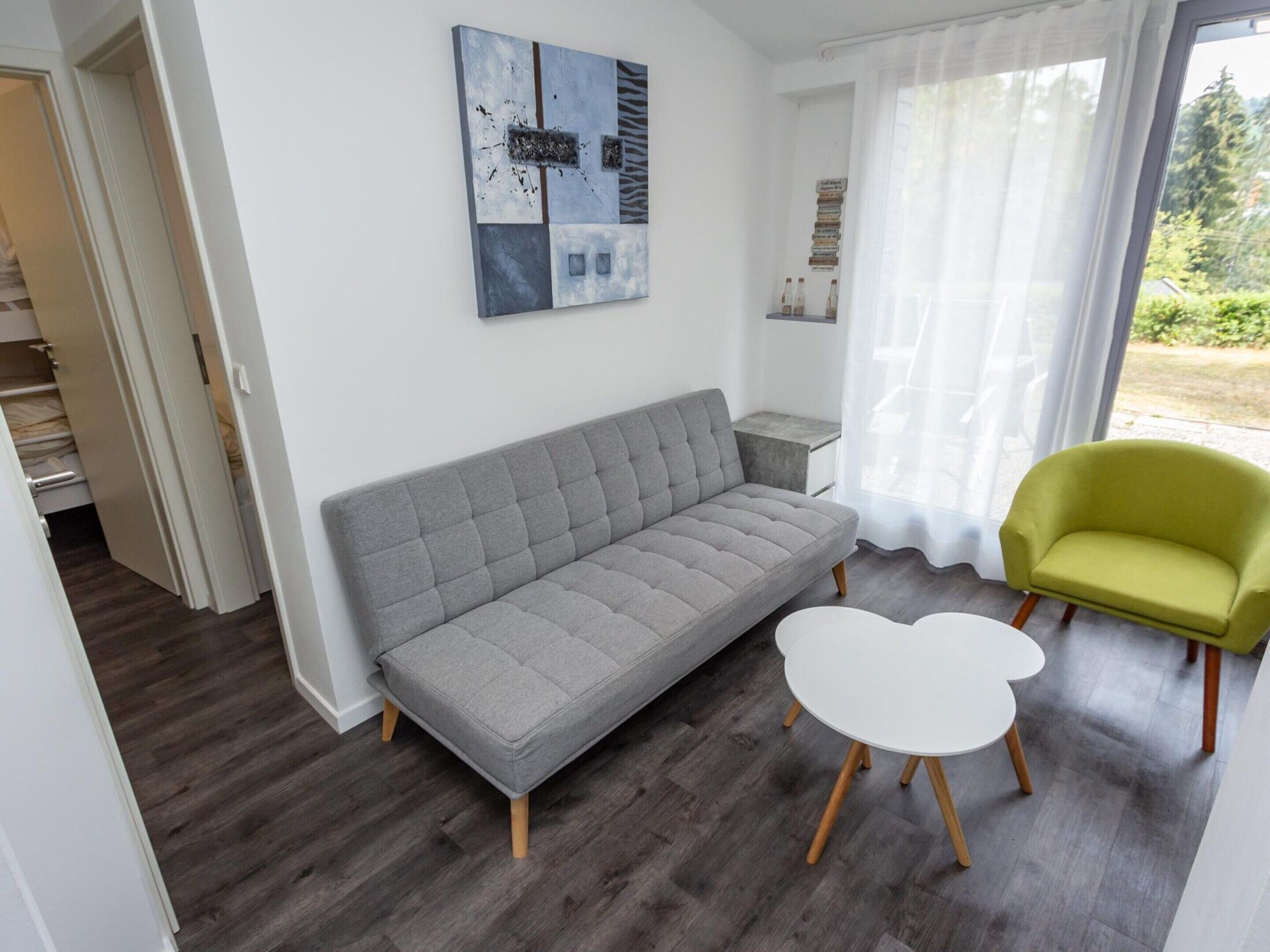 Una sala de estar moderna con un sofá gris, una mesa de centro blanca y un sillón verde. Hay un cuadro en la pared y una ventana con cortinas translúcidas.