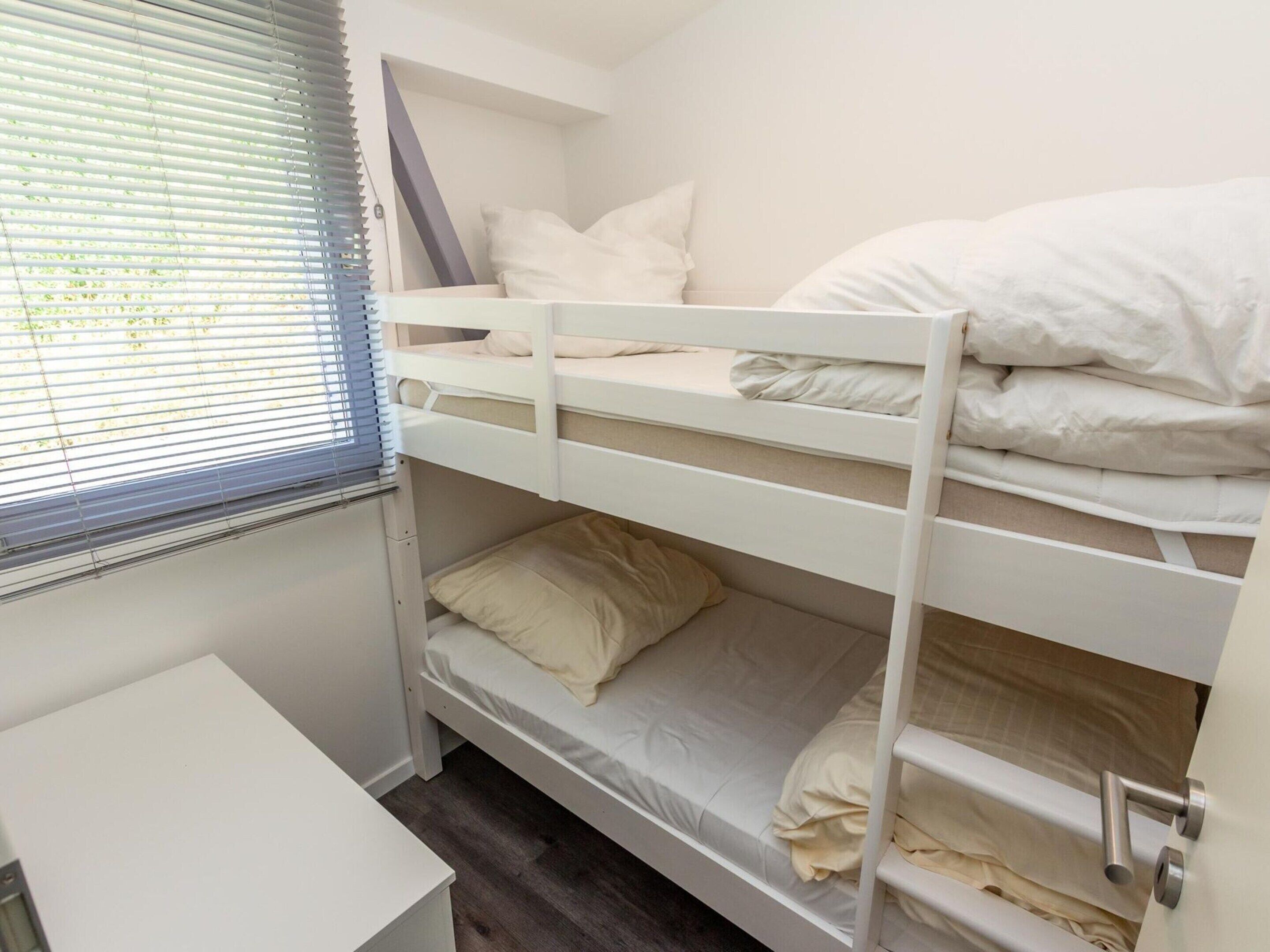 Une chambre avec un lit superposé, un bureau blanc et une fenêtre avec des stores.
