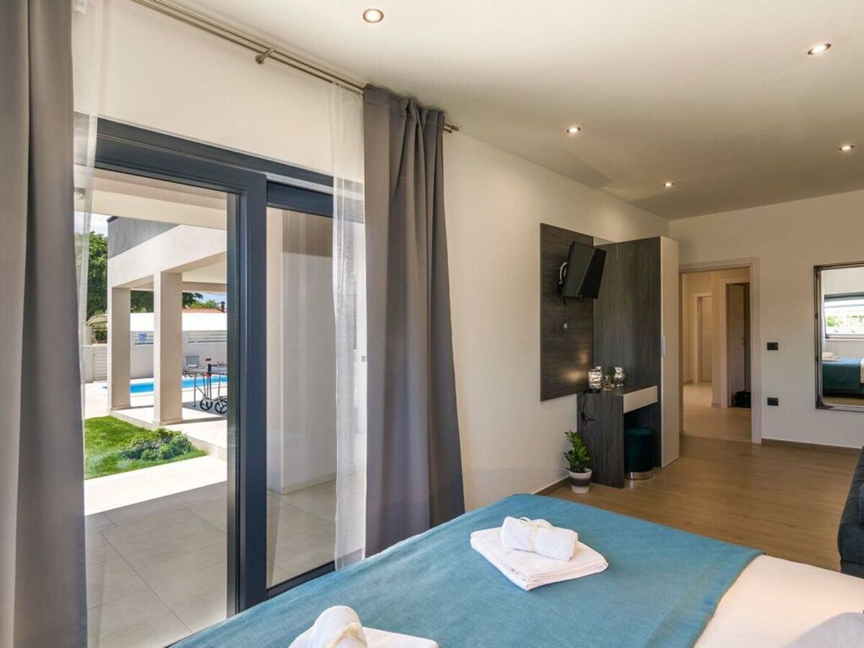 A modern hotel room with a large bed, a TV mounted on the wall, and a view of the pool outside.