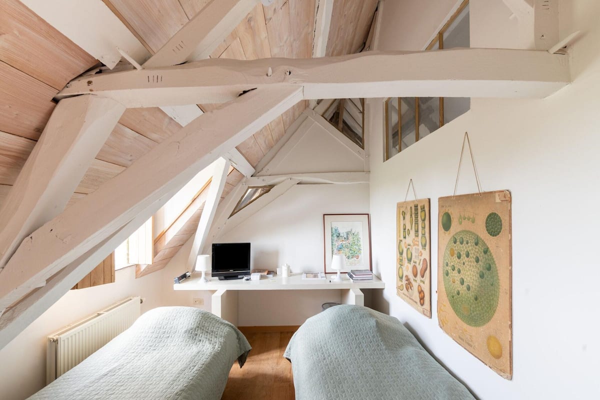 A cozy attic bedroom with exposed wooden beams, a desk with a computer, and two beds.