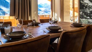 Een eetruimte met een houten tafel voor zes personen, grote ramen met uitzicht op de bergen, en een schilderij van een waterval aan de muur.