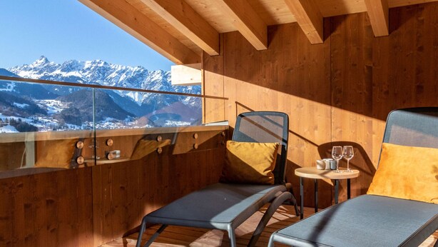 Een houten interieur met uitzicht op besneeuwde bergen. Er staan twee stoelen en een klein tafeltje met glazen bij.