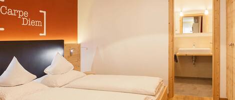 A hotel room with two beds, a wooden headboard, a wall-mounted light, and a wooden door leading to a bathroom with a sink and mirror.
