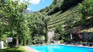 A swimming pool with lounge chairs and a table, surrounded by lush greenery and a mountainous backdrop. - Apartment Carpe Diem (Merano)