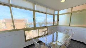 A dining area with a glass table and white chairs, large windows with a view of buildings and a body of water. - Belvilla by OYO Villa in La Manga del mar Menor (Cartagena)