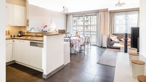 A modern apartment with a kitchen, dining area, and living room. It features a large window, a dining table with chairs, a sofa, and a TV. - Apartment by the Coast With Sunny Terrace (Koksijde)