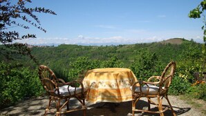 A table with a tablecloth and two wicker chairs on a stone surface, overlooking a mountainous landscape with greenery and a clear sky. - Holiday Home Cascina Lovera (Bonvicino)