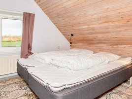 Un dormitorio con techo inclinado, una cama con ropa blanca, un ventanal con vistas a la vegetación y una alfombra estampada.