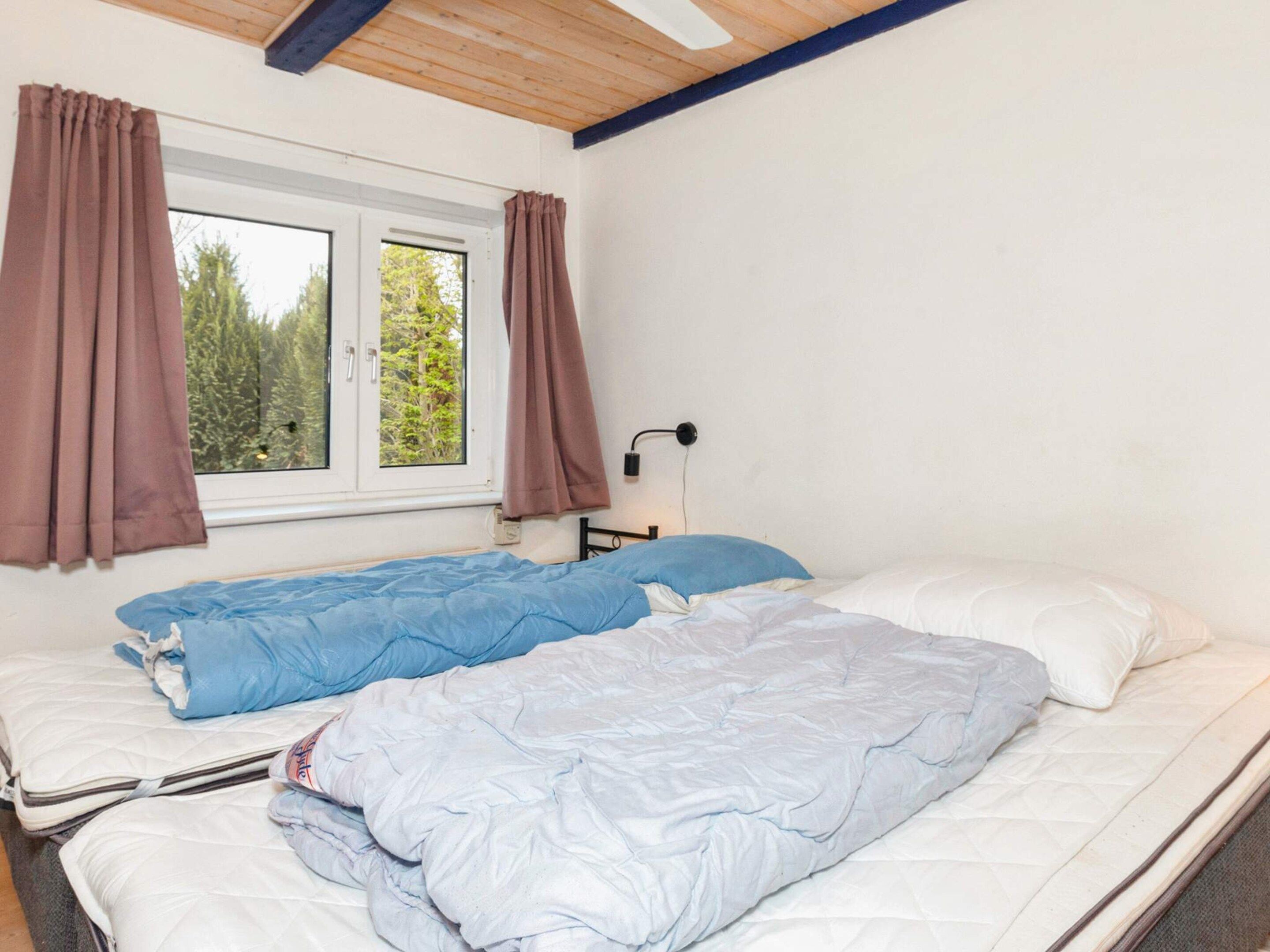 A room with two beds, a window with curtains, a wall-mounted lamp, and a view of trees outside.