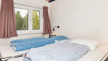 A room with two beds, a window with curtains, a wall-mounted lamp, and a view of trees outside.